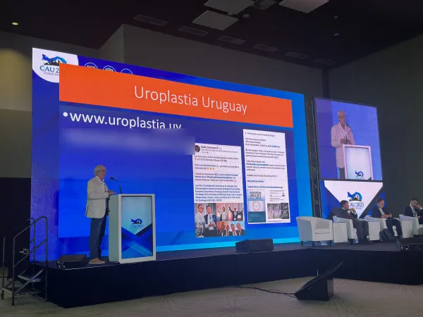 El Dr. Javier Zeballos exponiendo desde el atril en el escenario del CAU 2025, con una pantalla gigante que dice Uroplastia Uruguay